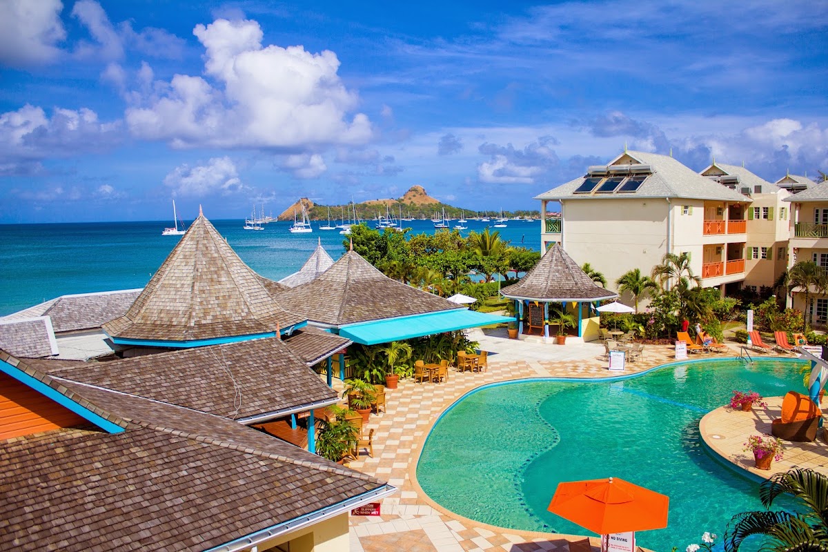 Resort pool with turquoise water and orange umbrellas, surrounded by cream-colored buildings with conical roofs, tropical landscaping, and sailboats anchored in the blue bay beyond.