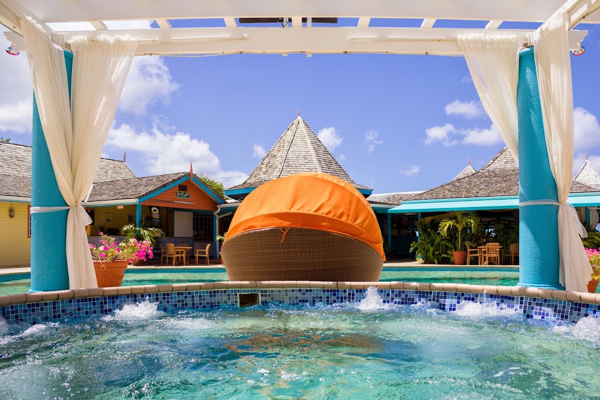Whirlpool spa with orange canopy in foreground, turquoise and yellow colonial-style buildings with conical roofs beyond, framed by white curtained cabana.