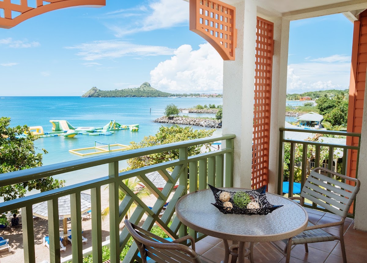 Beachfront balcony at Bay Gardens Beach Resort & Spa with round table, chairs, and view of turquoise water, inflatable water toys, and forested island beyond.