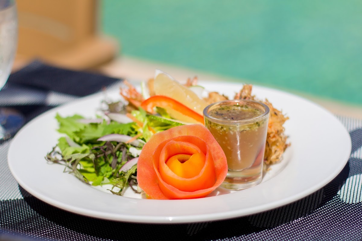 Plated dish with mixed greens, sliced red pepper rose, fried shrimp, and dressing served beachside with turquoise water in background.