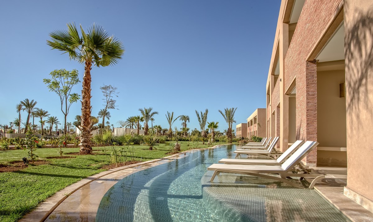 Lounge chairs line a narrow pool beside a brick resort building, with manicured lawns and palm trees extending across the grounds under clear blue sky.
