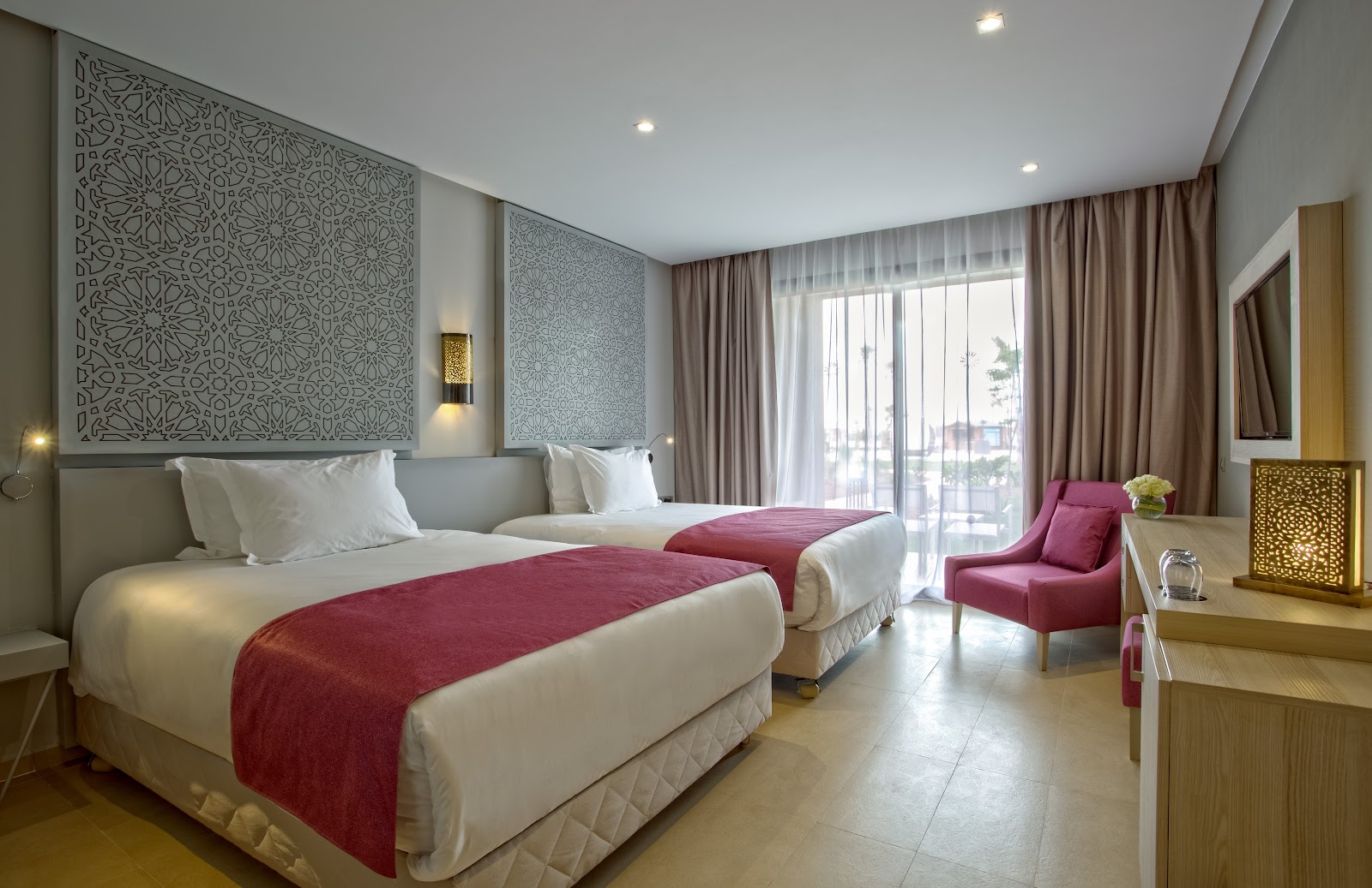 Hotel room with two queen beds dressed in white linens and red runners, geometric patterned headboards, magenta chair, and balcony overlooking palm trees.