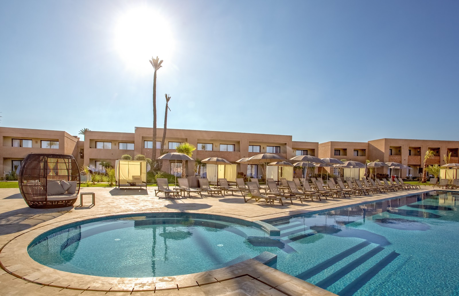Pool area with lounge chairs, umbrellas, and palm trees at Be Live Collection Marrakech Adults Only, with tan resort buildings in background.
