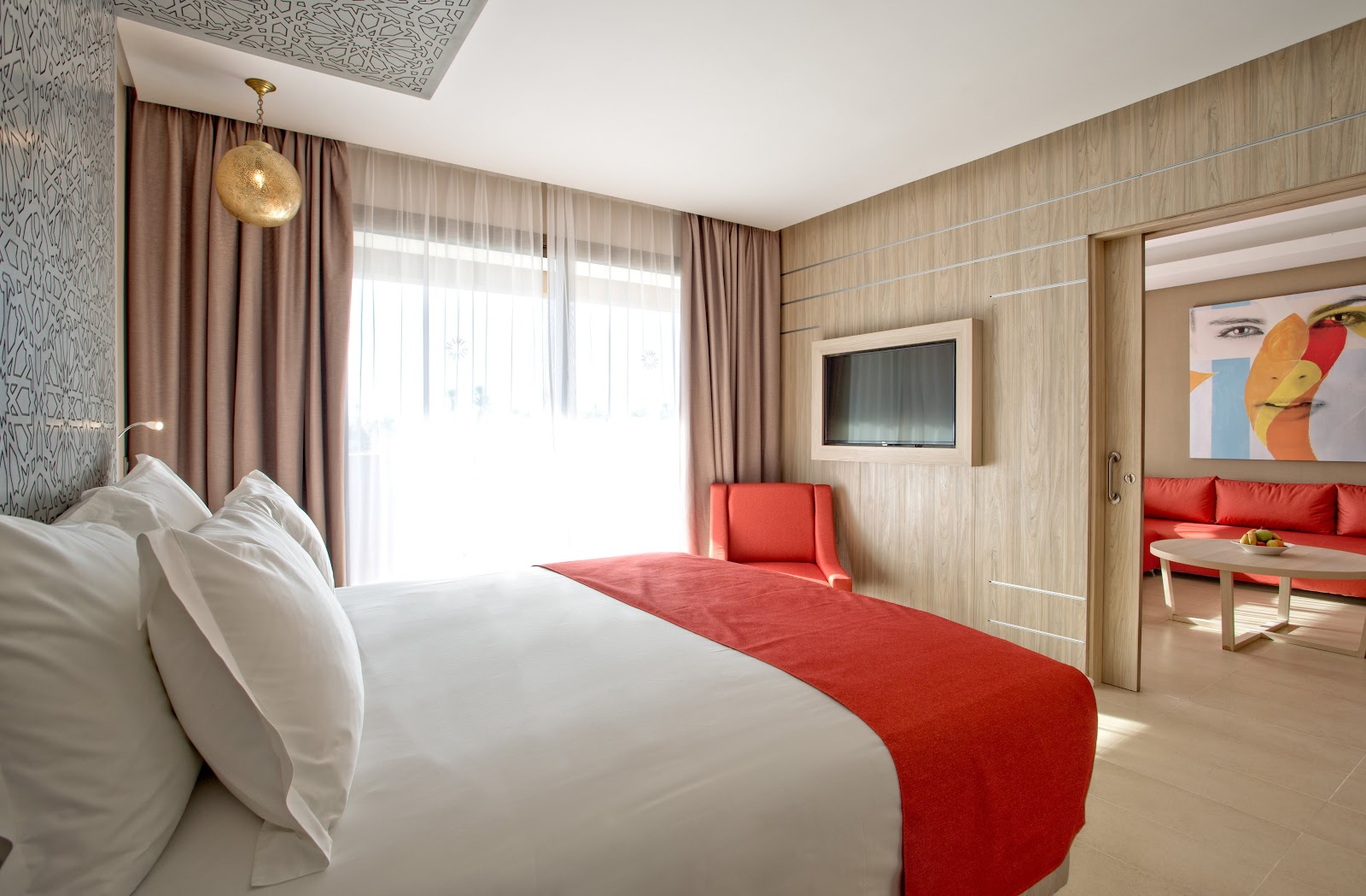 Modern hotel suite with white bedding, red accent runner, wood-paneled walls, and open doorway to living area with red sofa.
