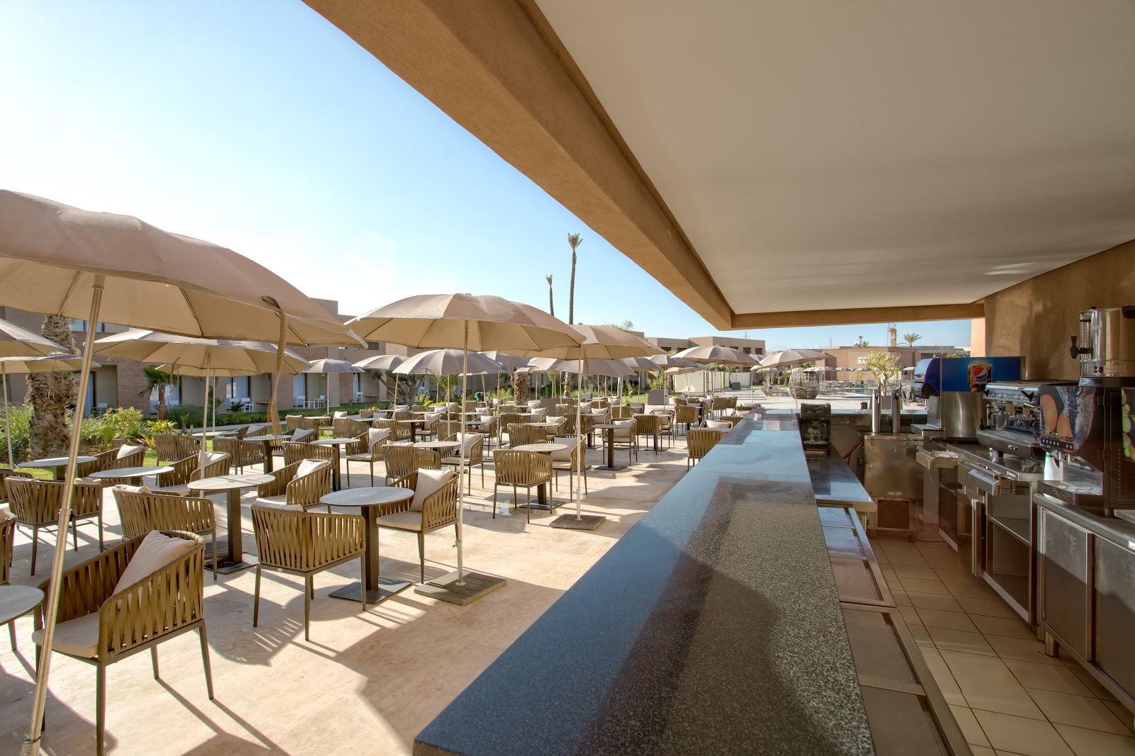 Covered dining area with tan umbrellas, woven chairs, and tables overlooking a sun-drenched courtyard at Be Live Collection Marrakech Adults Only.