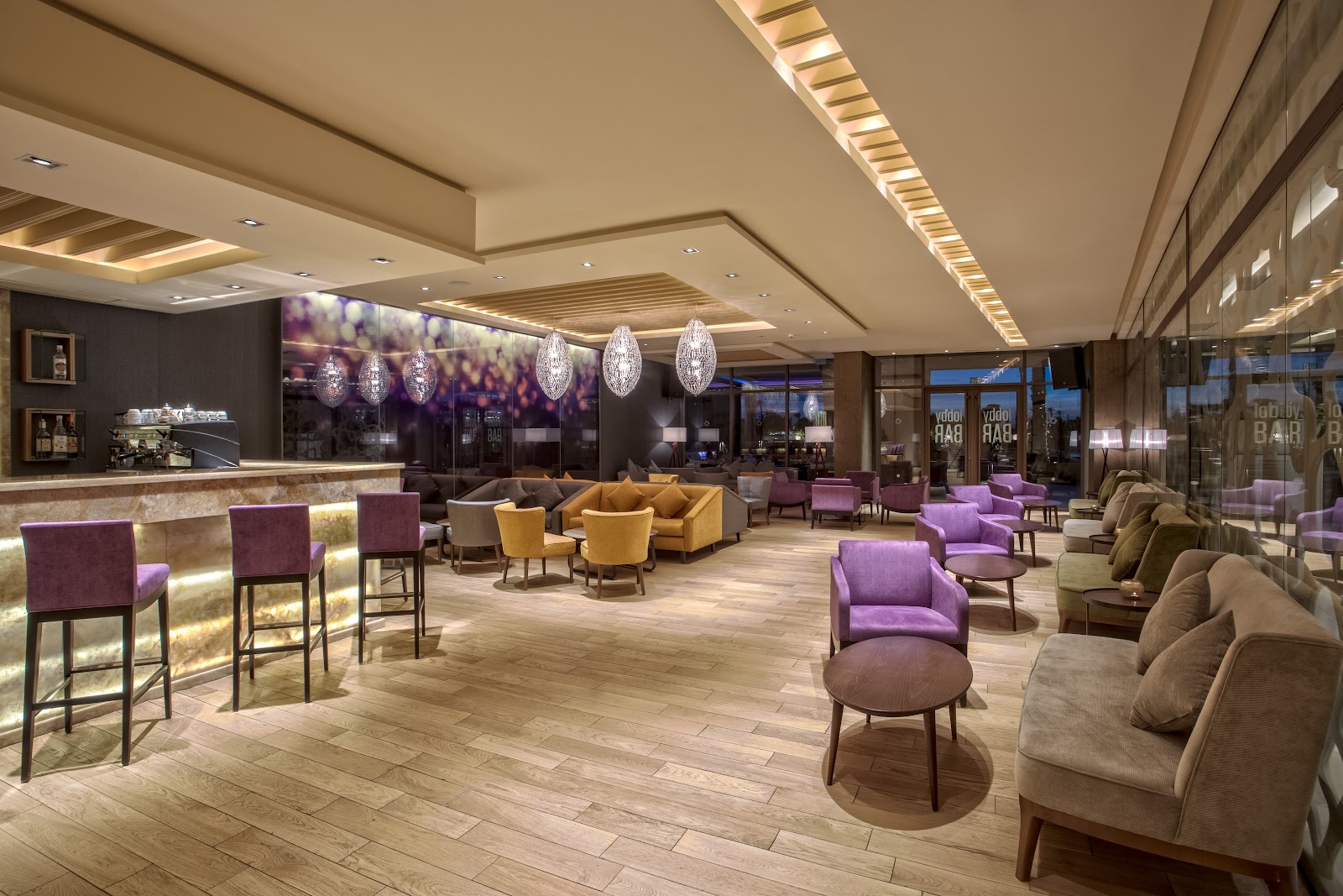 Modern lounge with purple and gold seating, illuminated bar, crystal pendant lights, and floor-to-ceiling windows overlooking evening cityscape.