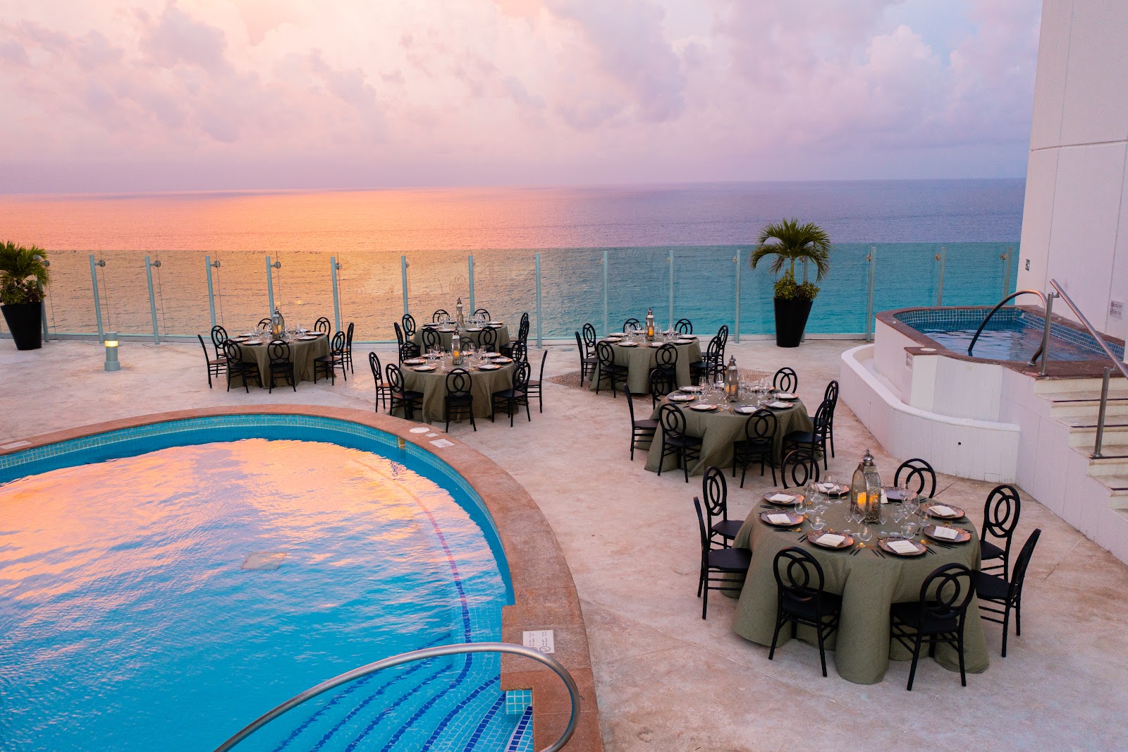 Beachfront dining terrace at Beach Palace with round tables set for dinner, pools, and turquoise ocean at sunset.