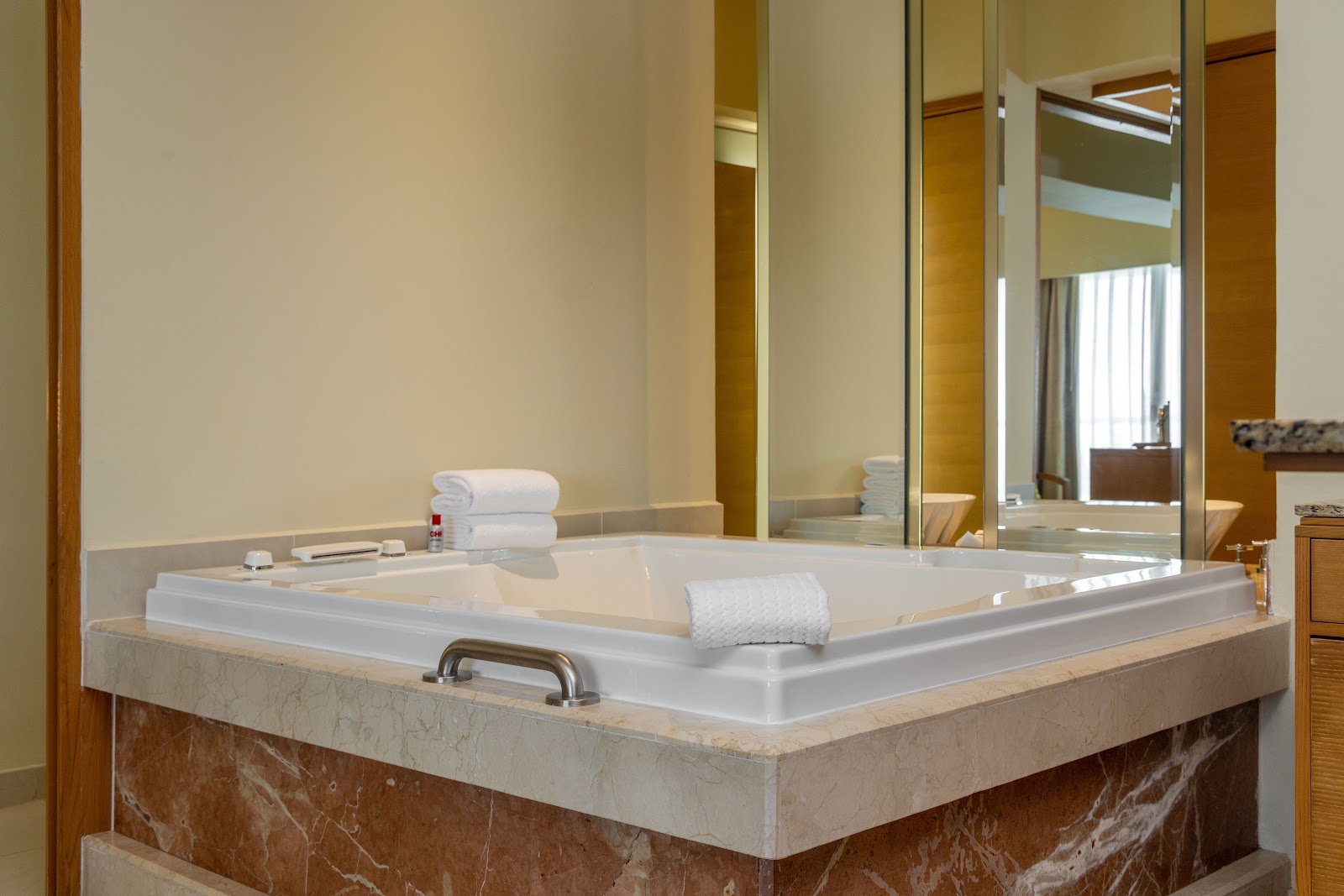 Resort bathroom with large soaking tub set in marble surround, white towels, and mirror reflecting bedroom beyond.