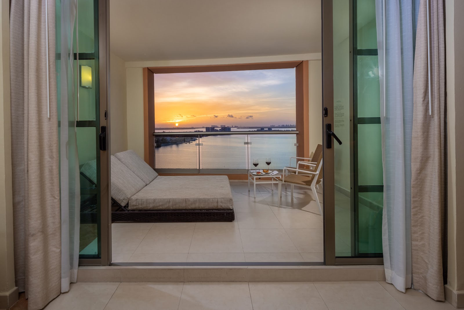 Modern beachfront suite with open doors framing a sunset view over calm water, lounge chair, and seating area on the balcony.