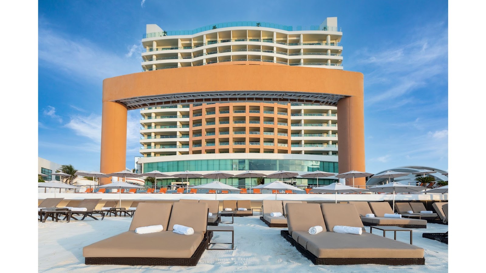 Lounge chairs line a sandy beach facing the curved orange and cream architecture of Beach Palace resort under blue sky.
