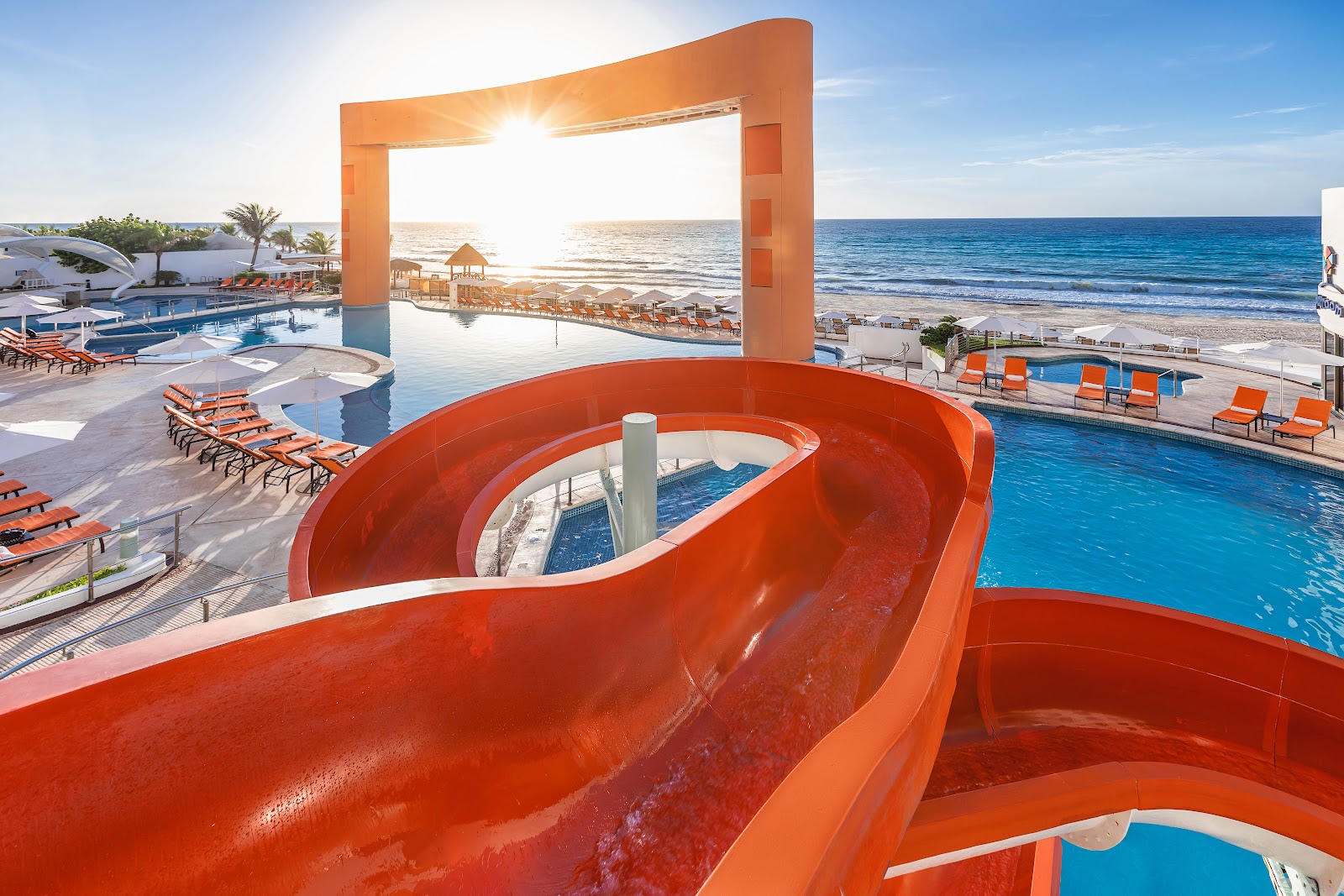 Red water slide curves over pools and beach with ocean and lounge chairs beyond at beachfront resort.