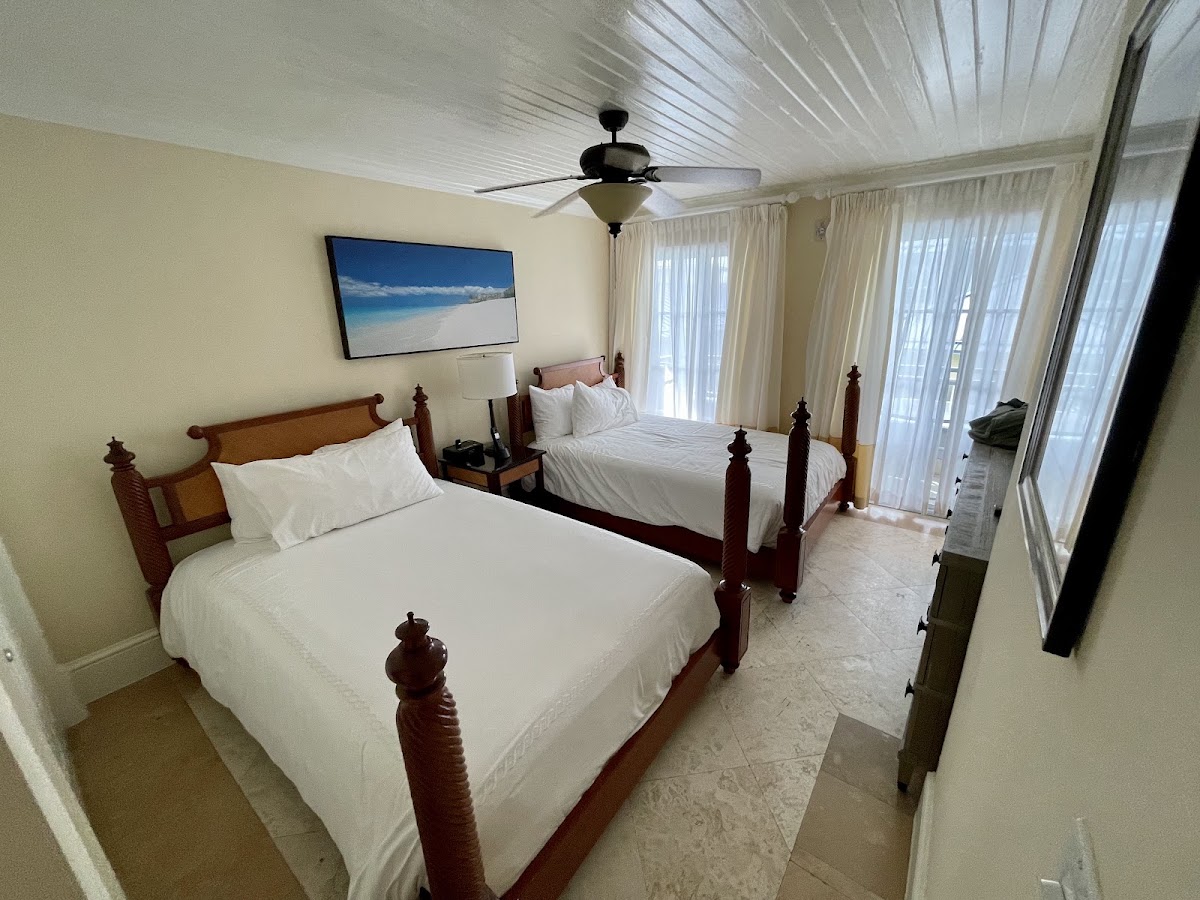 Resort room with two wooden poster beds with white linens, cream walls, ceiling fan, TV displaying a beach scene, and glass doors opening to a balcony.
