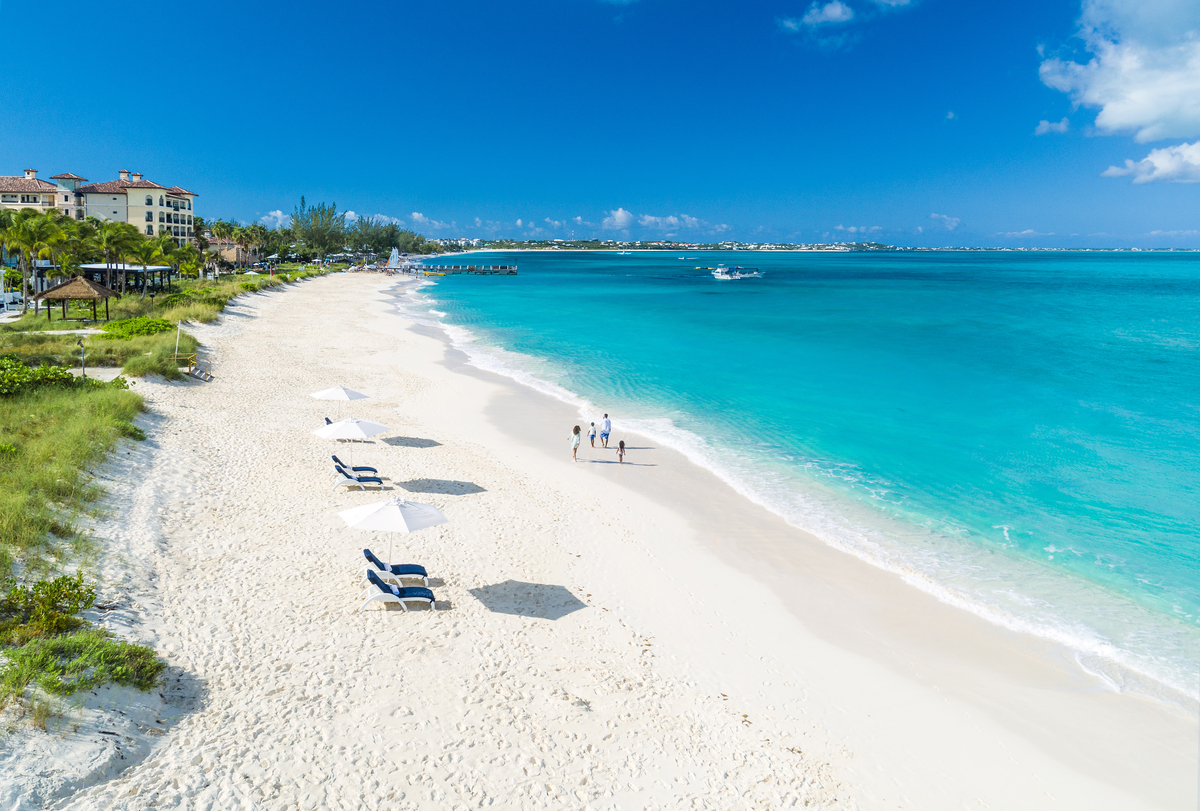 White sand beach with turquoise water, lounge chairs, guests wading, resort buildings and palm trees along the shoreline.
