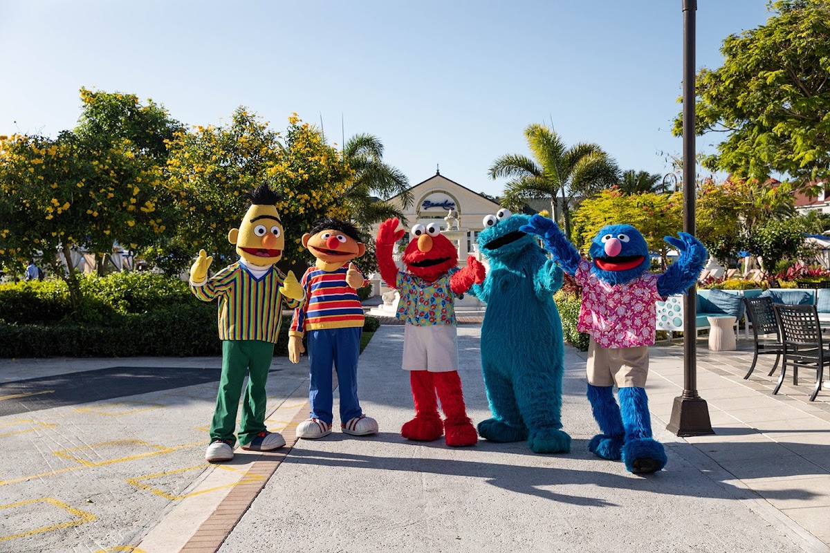 Sesame Street characters stand on a paved plaza at Beaches Turks & Caicos surrounded by tropical flowers and palm trees.