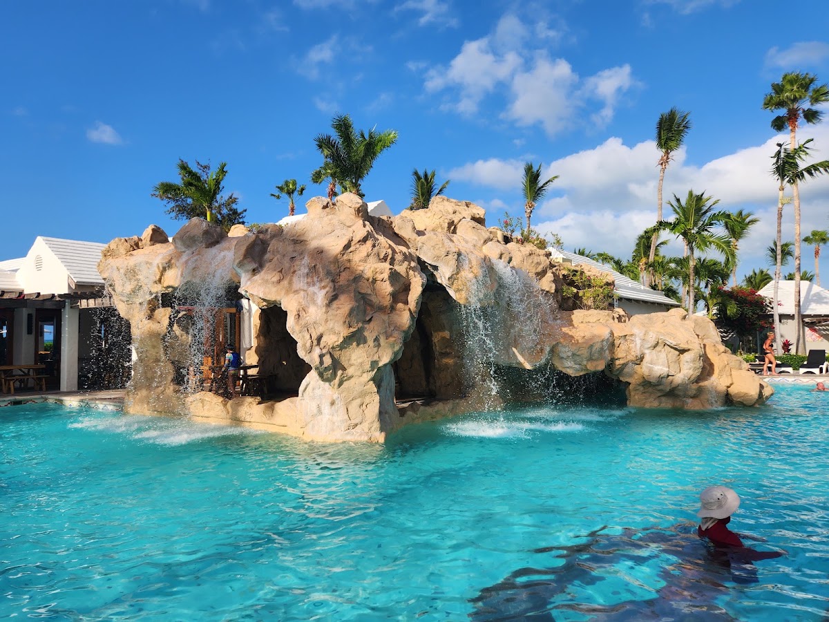 Turquoise pool with large rock formation featuring waterfalls, palm trees, and white resort buildings under blue sky.