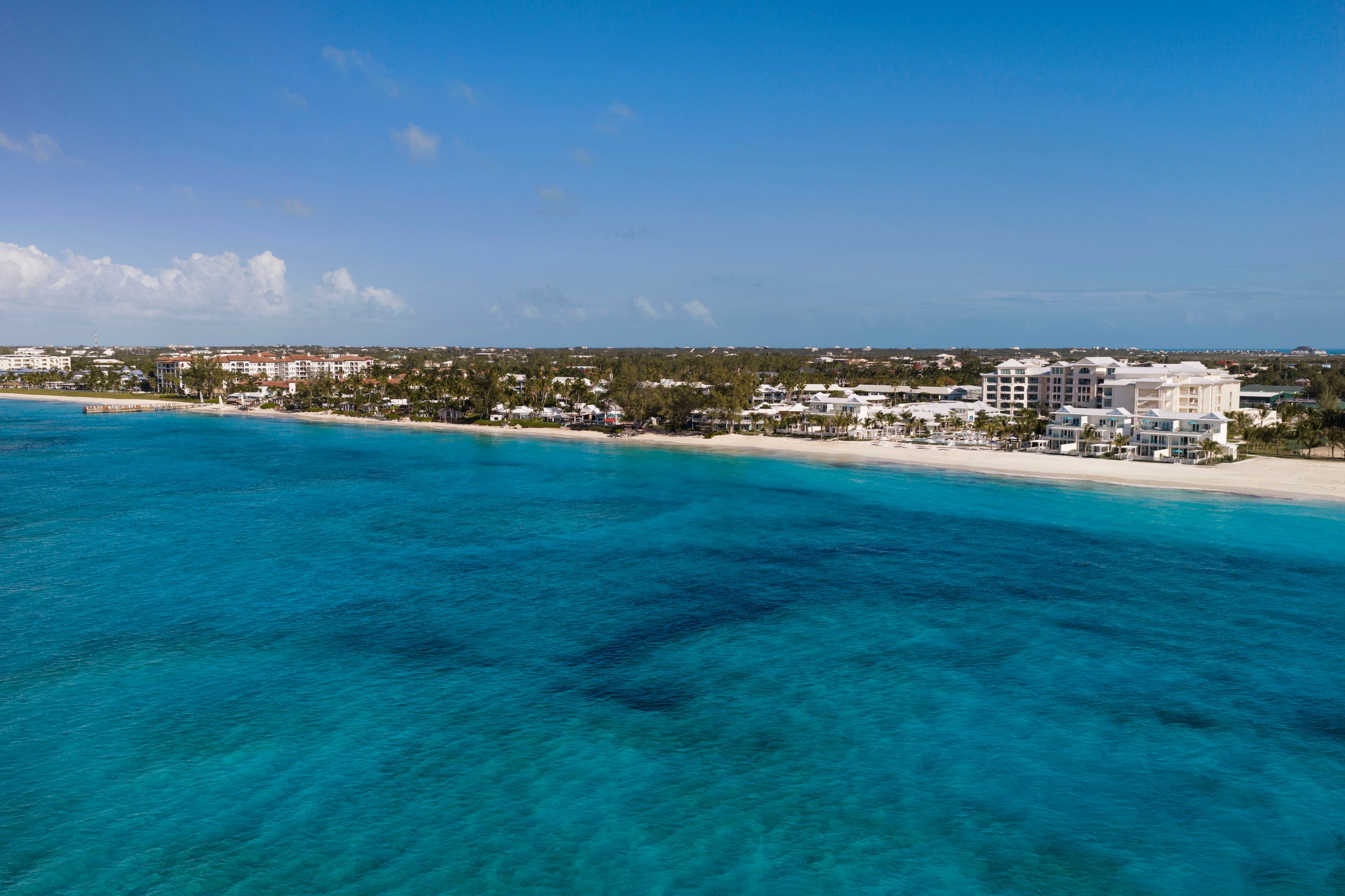 Low aerial over deep turquoise water toward the Treasure Beach Village shoreline.