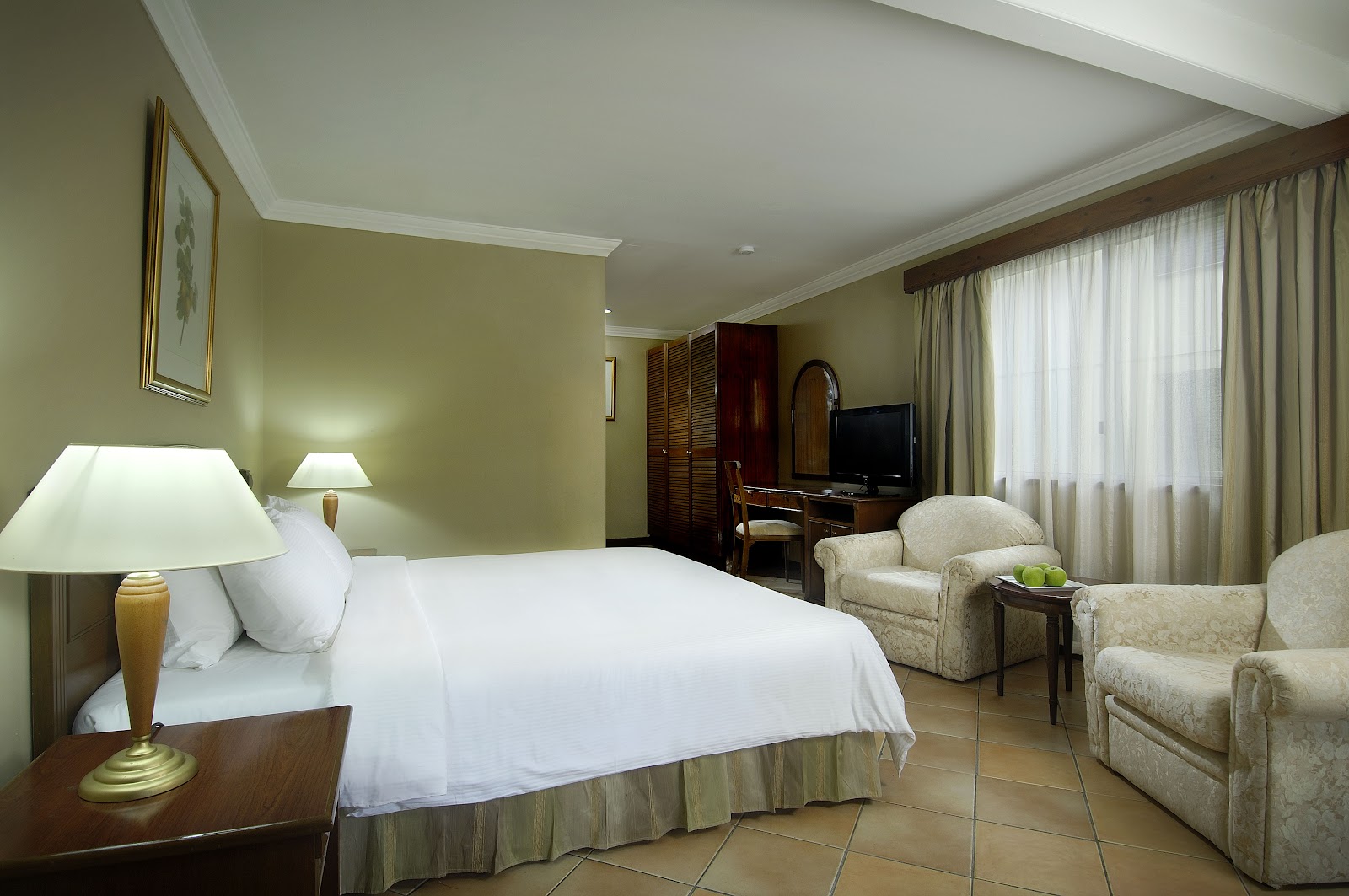 Resort guest room with queen bed, armchair, desk with flat-screen TV, and balcony overlooking tropical greenery.