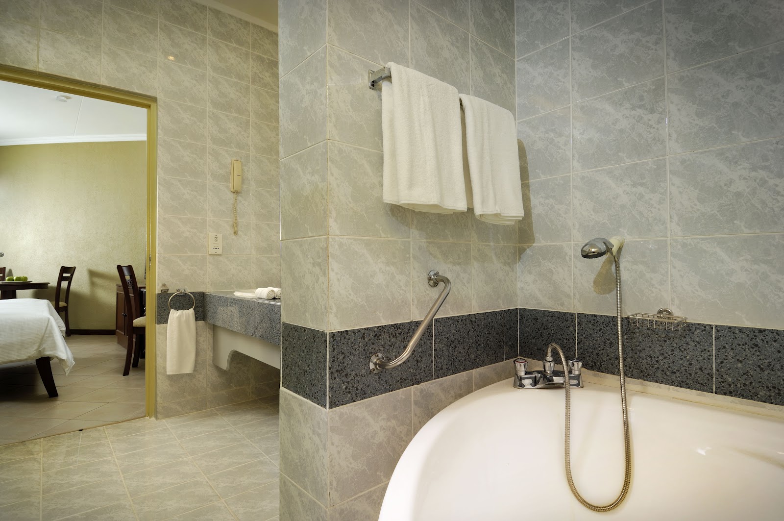 Resort bathroom with soaking tub, gray and beige tile walls, handheld showerhead, and white towels on rack, open to bedroom beyond.