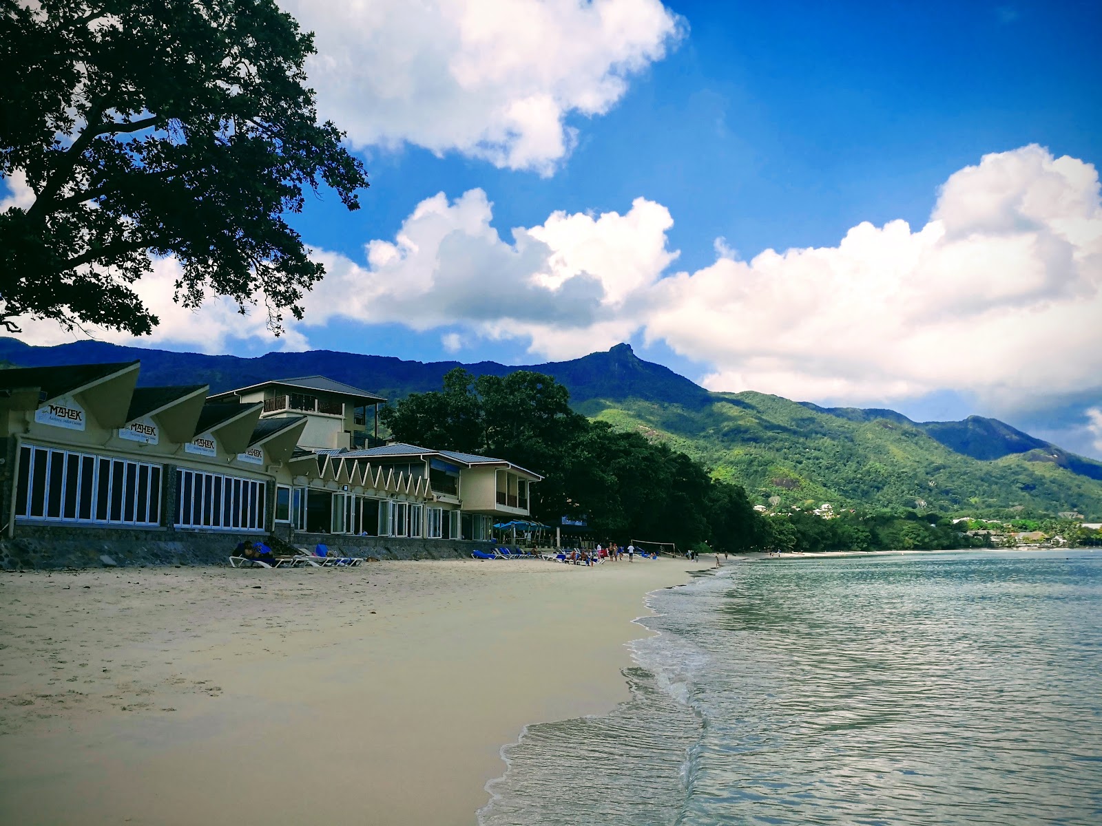 Resort pool with blue umbrellas and loungers overlooking forested mountains under partly cloudy sky.