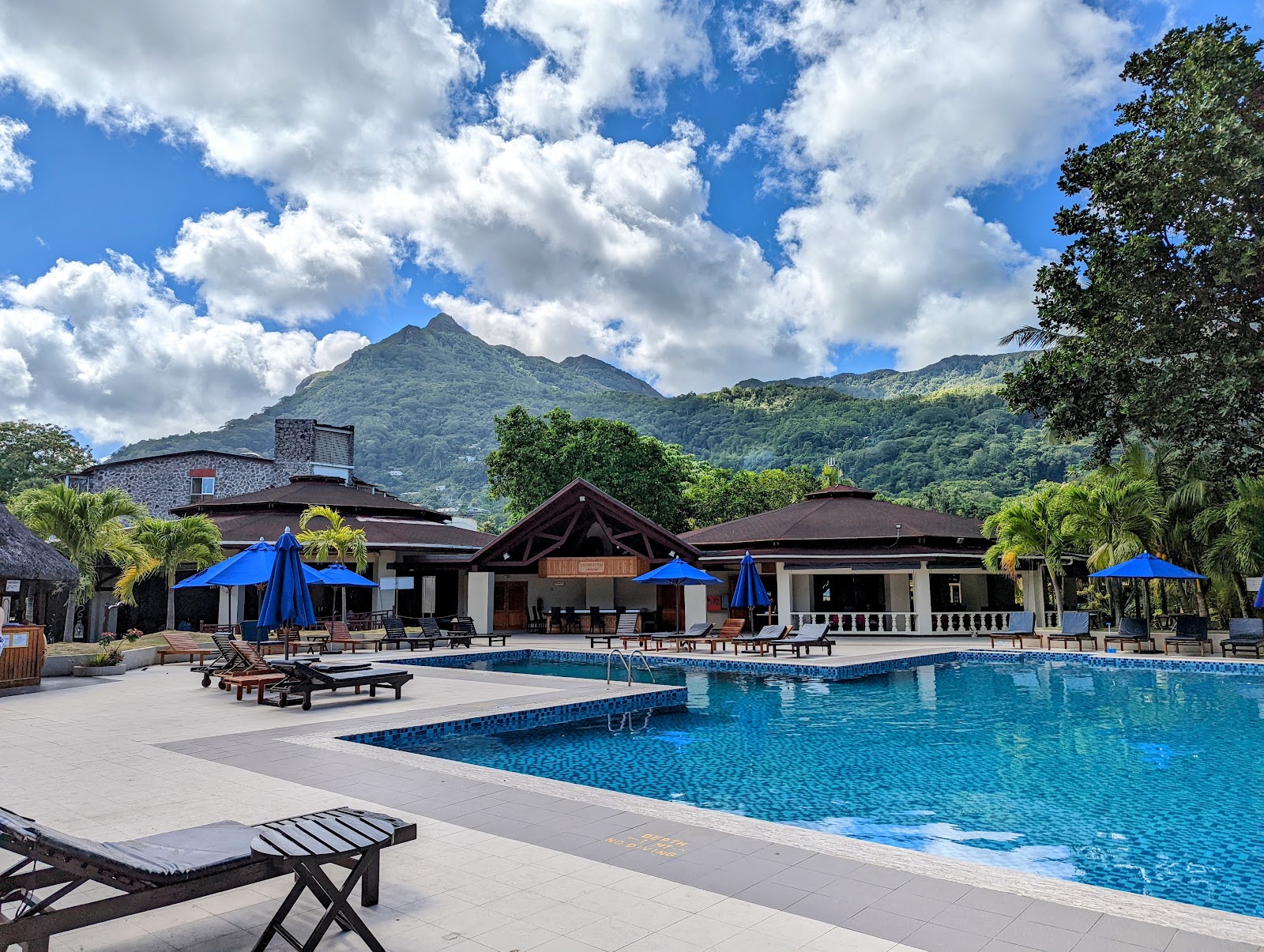Resort grounds with a central fountain surrounded by manicured lawn, palm trees, and colorful planted beds, with multi-story accommodation buildings and green mountains beyond.