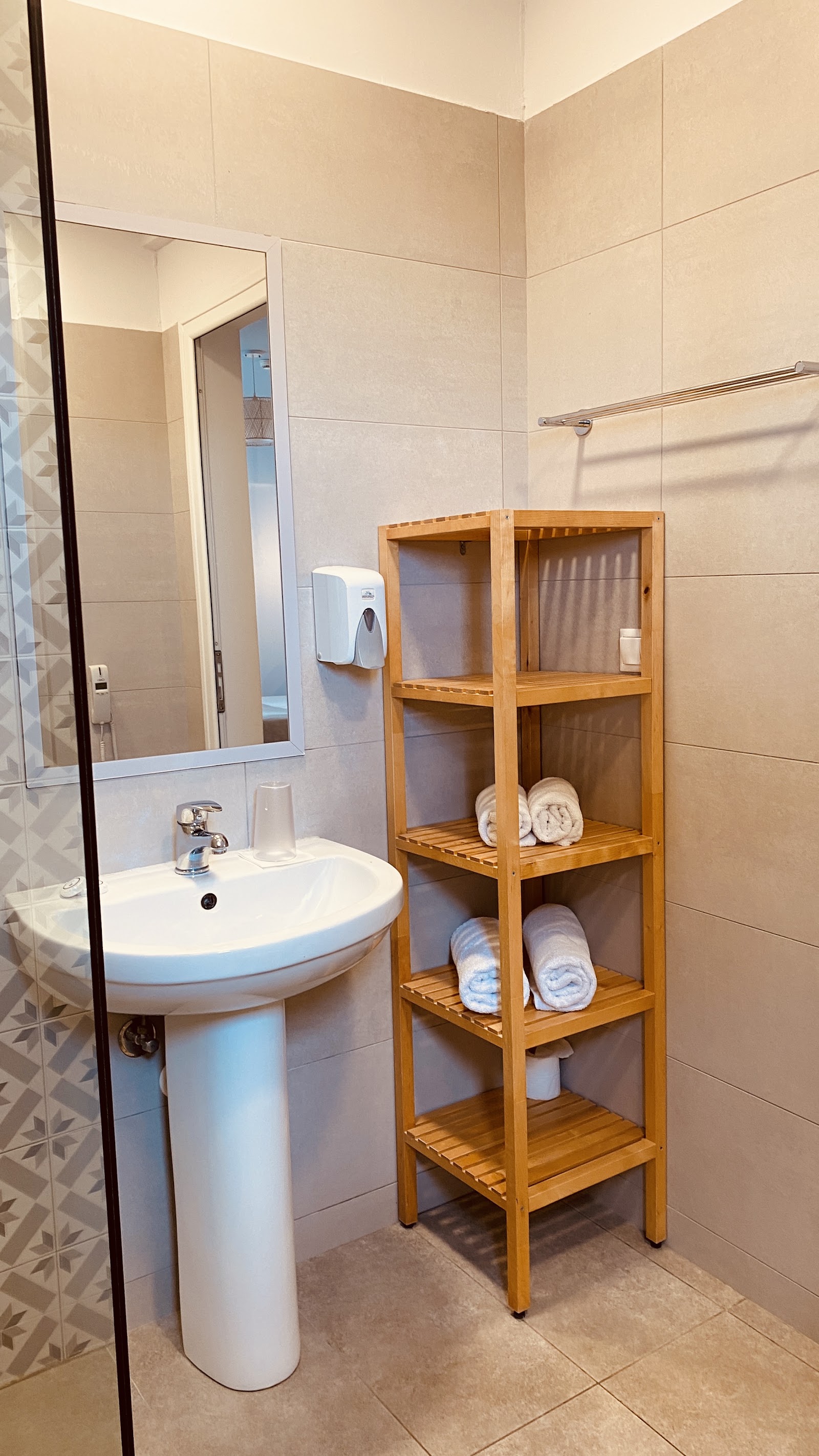 Resort bathroom with pedestal sink, mirror, wooden shelving unit holding rolled towels, and glass shower enclosure.