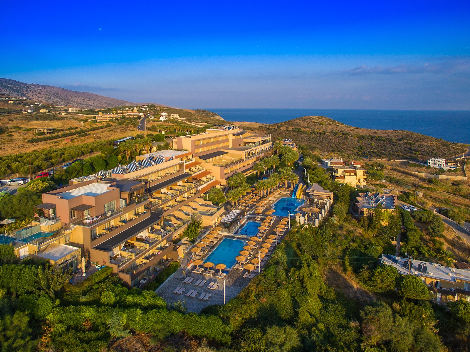 Resort with terracotta buildings nestled on a hillside above the Mediterranean Sea, featuring multiple pools surrounded by loungers and Mediterranean vegetation.