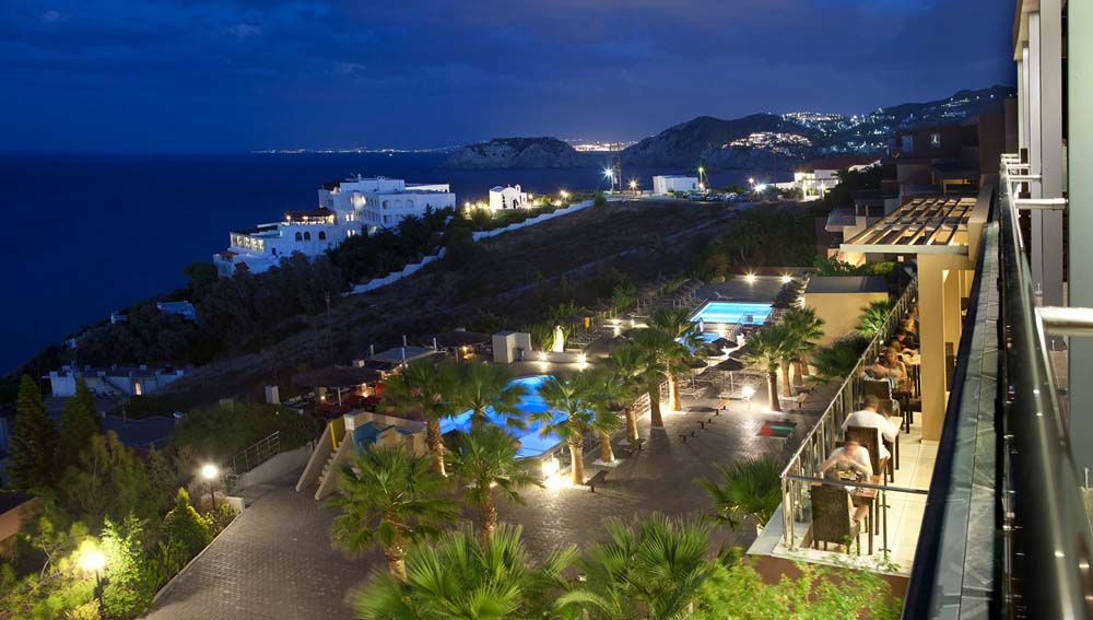Nighttime aerial view of Blue Bay Resort Hotel with illuminated pools, palm trees, and whitewashed buildings overlooking the dark Aegean Sea.