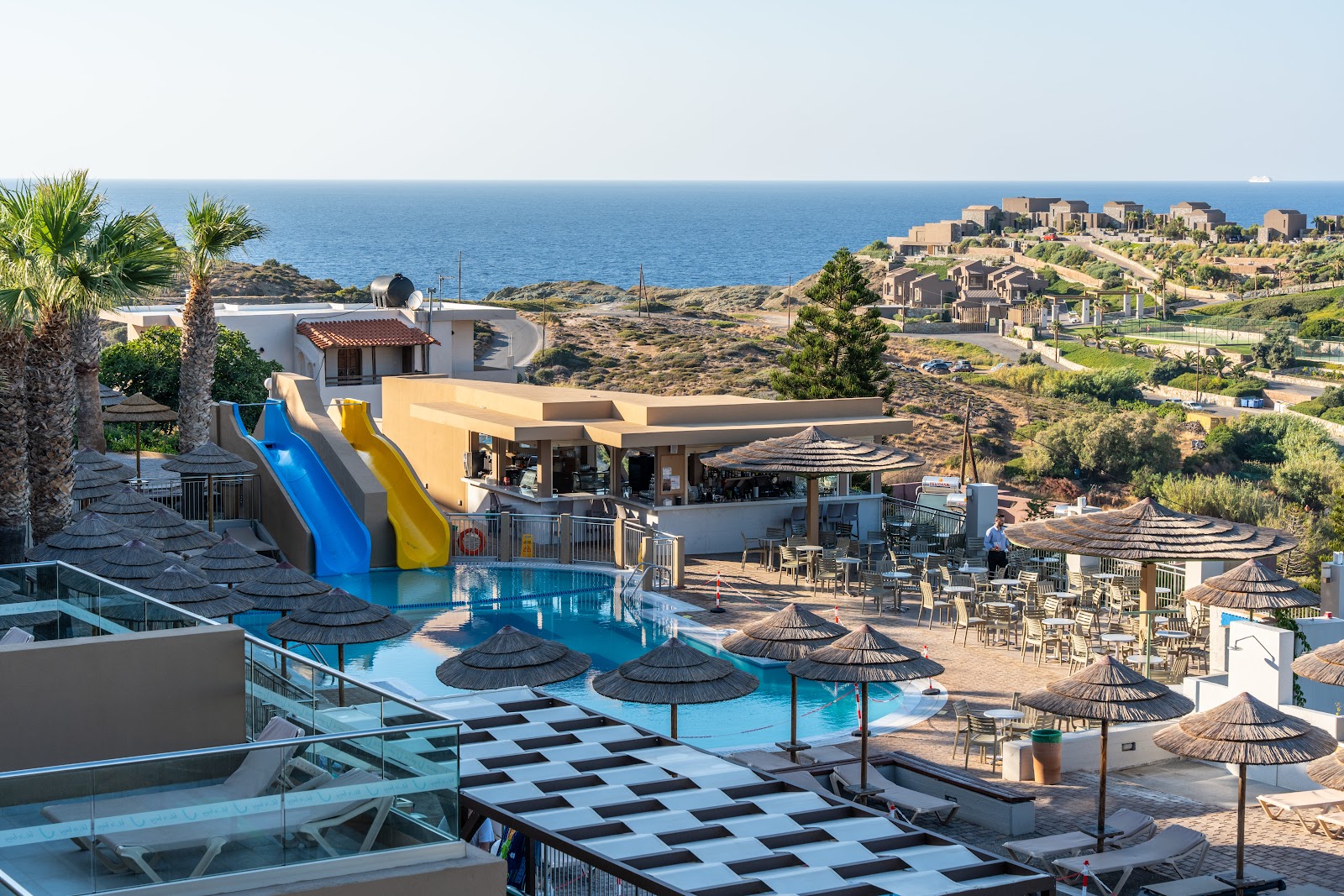 Blue Bay Resort Hotel's pool complex with blue and yellow water slides, thatched umbrellas, and Mediterranean coastline beyond.