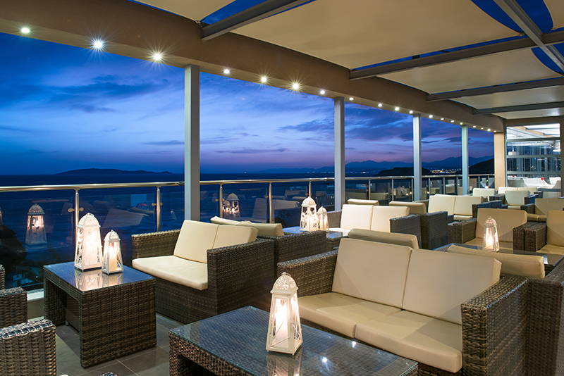Covered terrace lounge with woven furniture and cream cushions overlooking a twilight seascape and distant mountains.