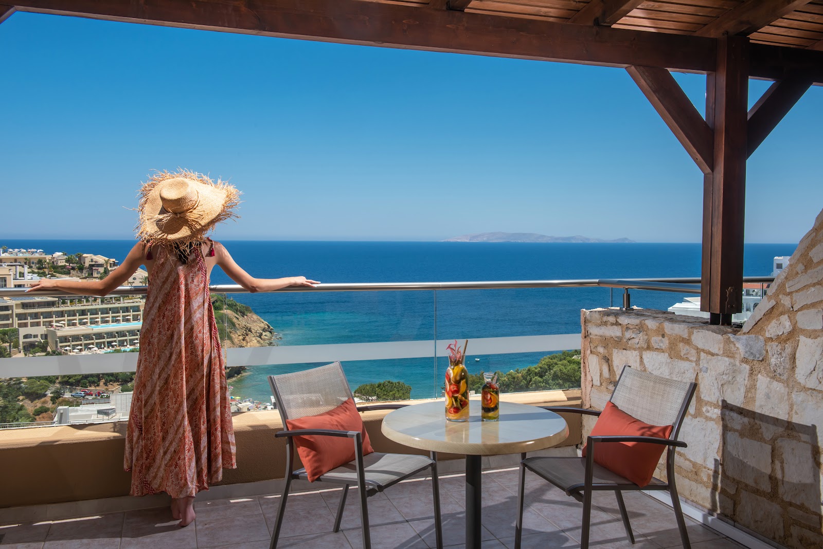 Woman in a straw hat stands on a cliffside terrace overlooking turquoise Mediterranean waters and distant coastline at Blue Bay Resort Hotel.