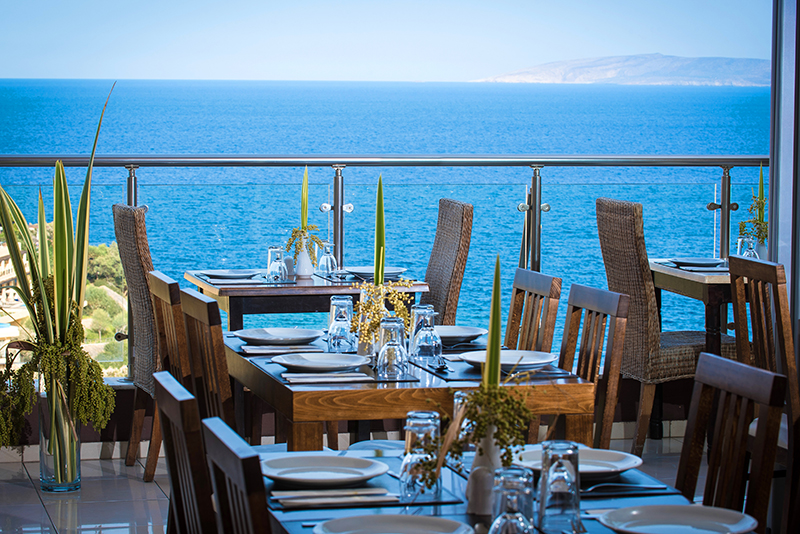 Dining room with wooden tables and chairs overlooking the blue sea through large windows at Blue Bay Resort Hotel.