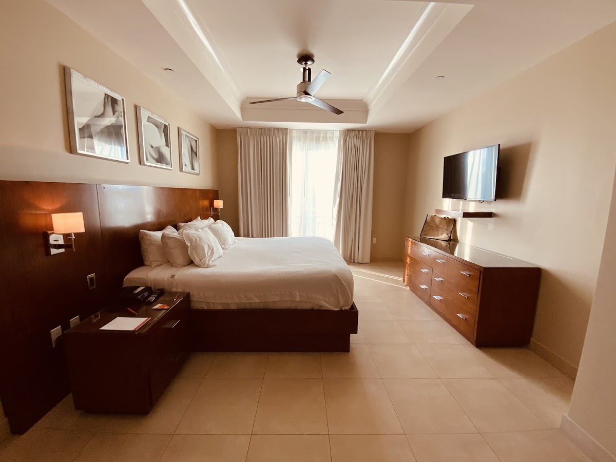 Resort bedroom with queen bed, dark wood headboard, white bedding, TV, dresser, and sliding glass doors to balcony with beige curtains.