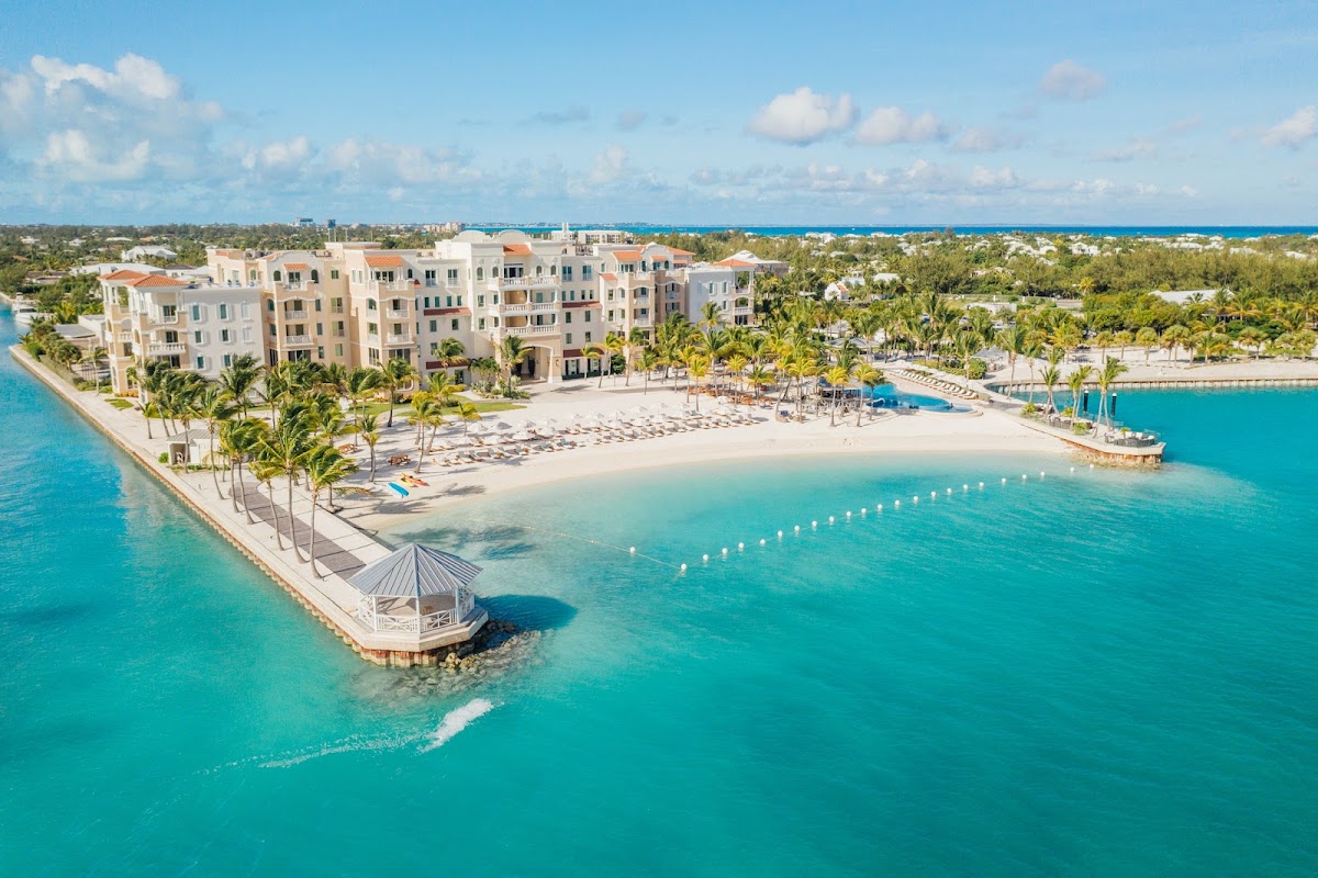 Blue Haven Resort beachfront with multi-story pastel building, white sand beach, lounge chairs, dock, and turquoise waters under clear sky.