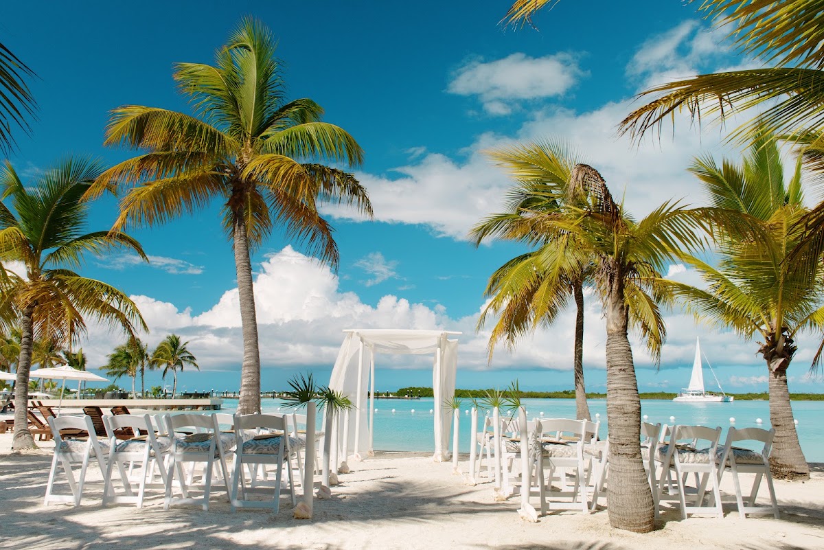 White wedding arbor and chairs set up on sandy beach at Blue Haven Resort with palm trees and turquoise water.