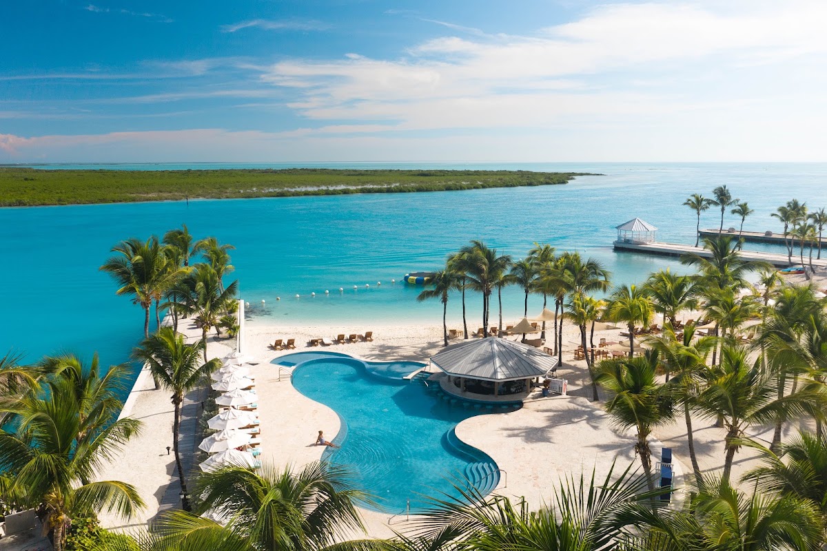 Blue Haven Resort's curved pool with thatched bar overlooks a turquoise lagoon, white sand beach, and mangrove coastline under clear sky.