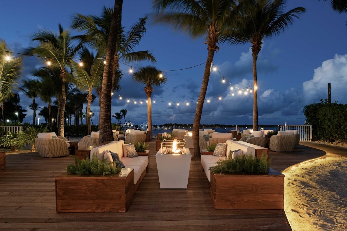 Blue Haven Resort's beachfront lounge at dusk with woven seating, palm trees, string lights, and a central fire pit overlooking the water.