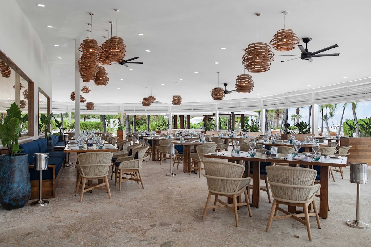 Dining room with woven pendant lights, wooden tables, wicker chairs, and floor-to-ceiling windows overlooking palm trees and beach at Blue Haven Resort.