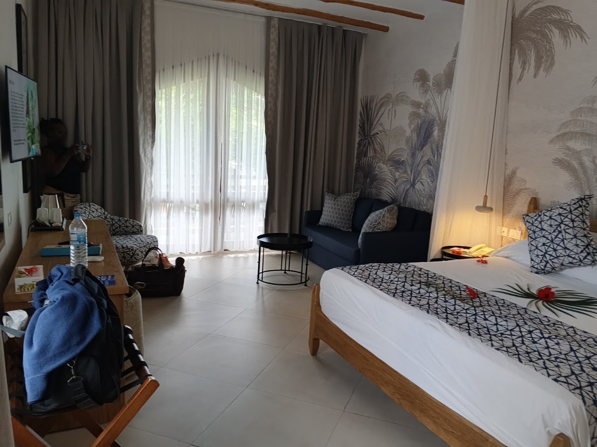 Resort guest room with bed, sofa seating area, tropical palm wallpaper, and glass doors opening to balcony at Bluebay Beach Resort & Spa.