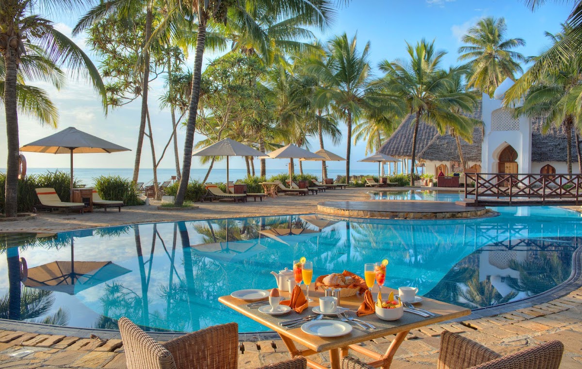 Breakfast table set poolside at Bluebay Beach Resort & Spa, with curved pool, palm trees, ocean view, and whitewashed architecture beyond.