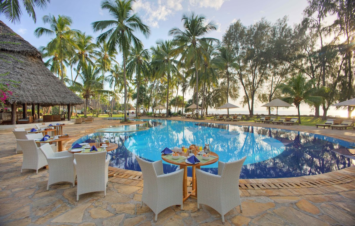 Bluebay Beach Resort & Spa pool area with white wicker chairs, round dining tables, and a large blue swimming pool surrounded by palm and acacia trees.