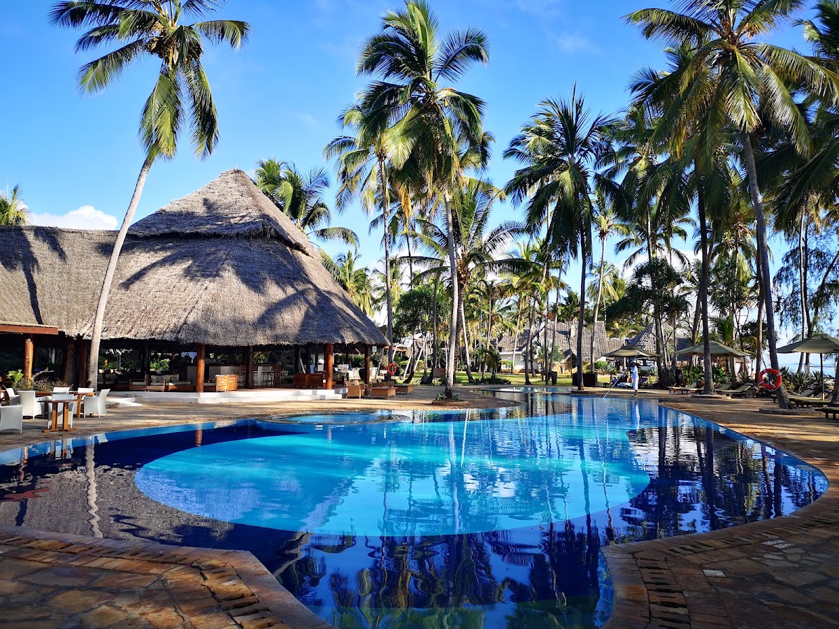 Bright blue pool surrounded by palm trees with thatched-roof lodge and wooden structures at Bluebay Beach Resort & Spa.