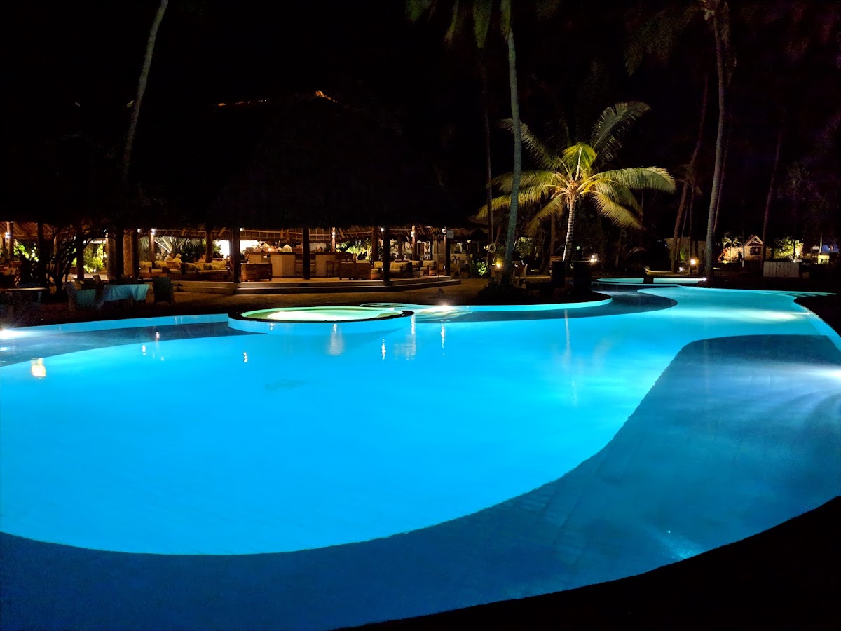 Illuminated curved pool at night with palm trees, lit tiki bar, and resort buildings in the background.