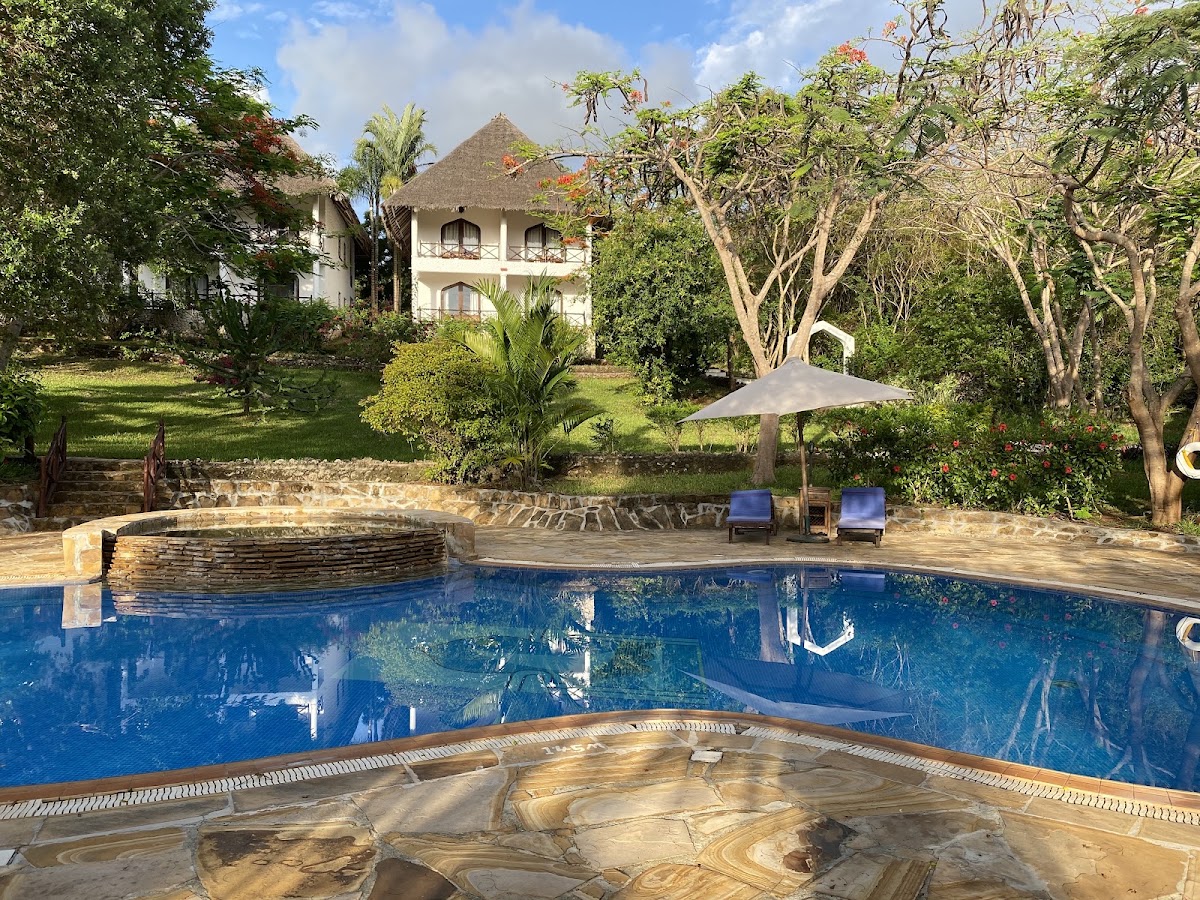 Blue infinity pool with stone deck facing a white two-story lodge with thatched roof, surrounded by tropical gardens and tall trees.