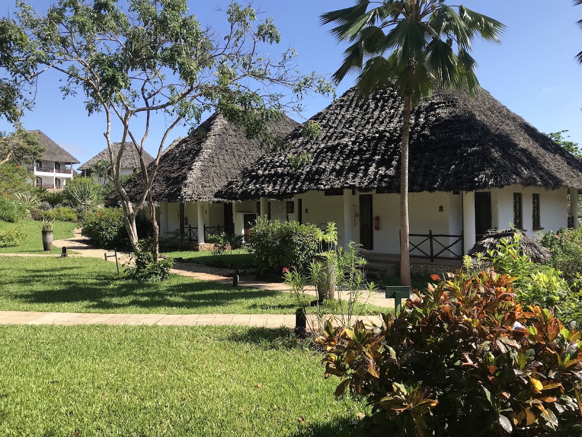 Thatched-roof bungalows with white walls and wooden verandas at Bluebay Beach Resort & Spa, surrounded by manicured lawns and tropical vegetation.