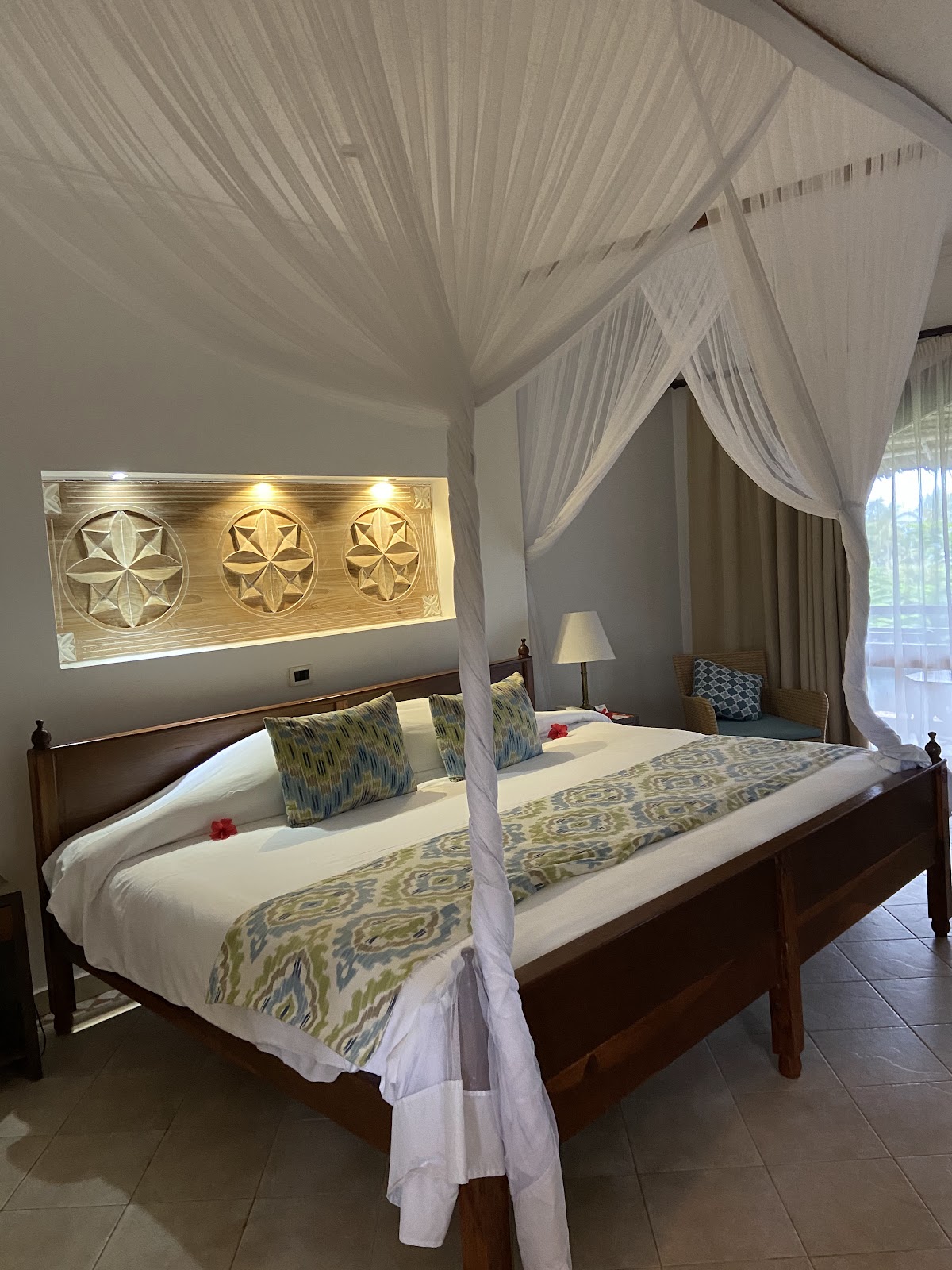 Four-poster bed with sheer white canopy drapes in a resort bedroom with geometric wall art and ocean view doors.