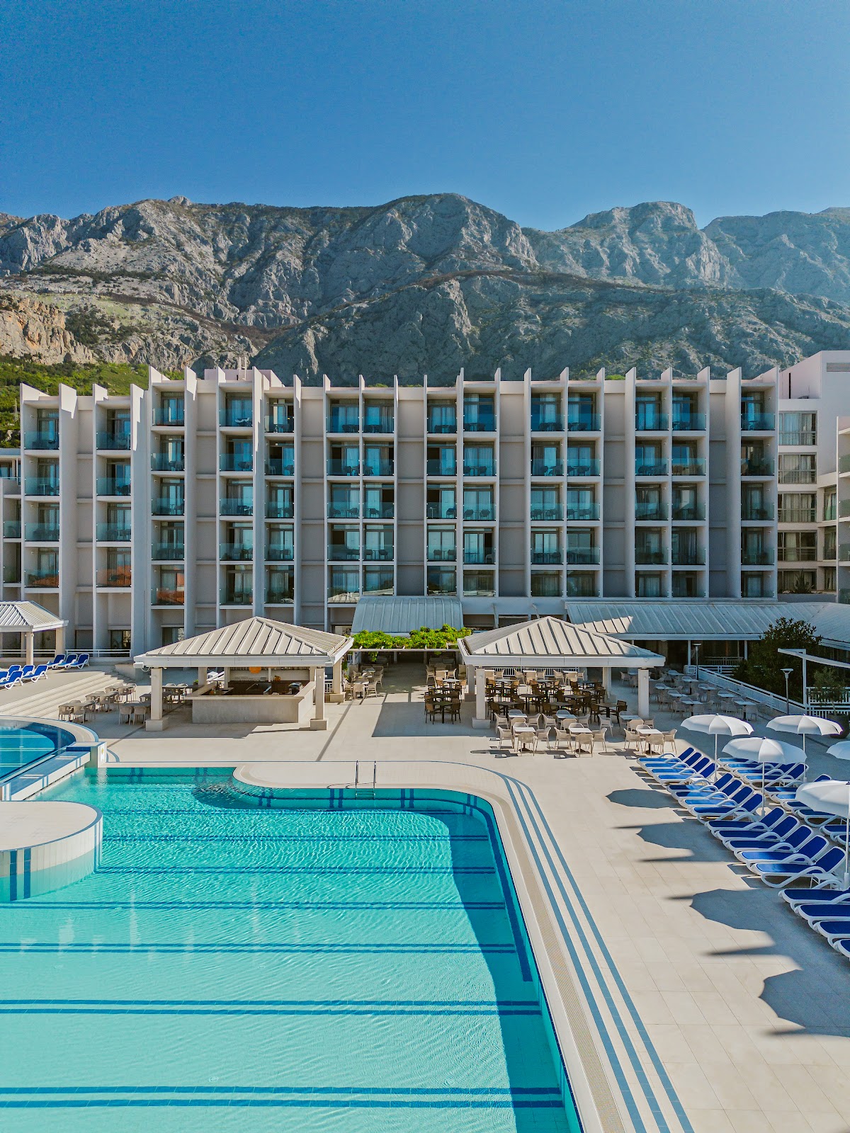 Modern white resort building with blue-windowed facade overlooking pool deck, loungers, and pergola-covered dining area beneath rocky mountains.