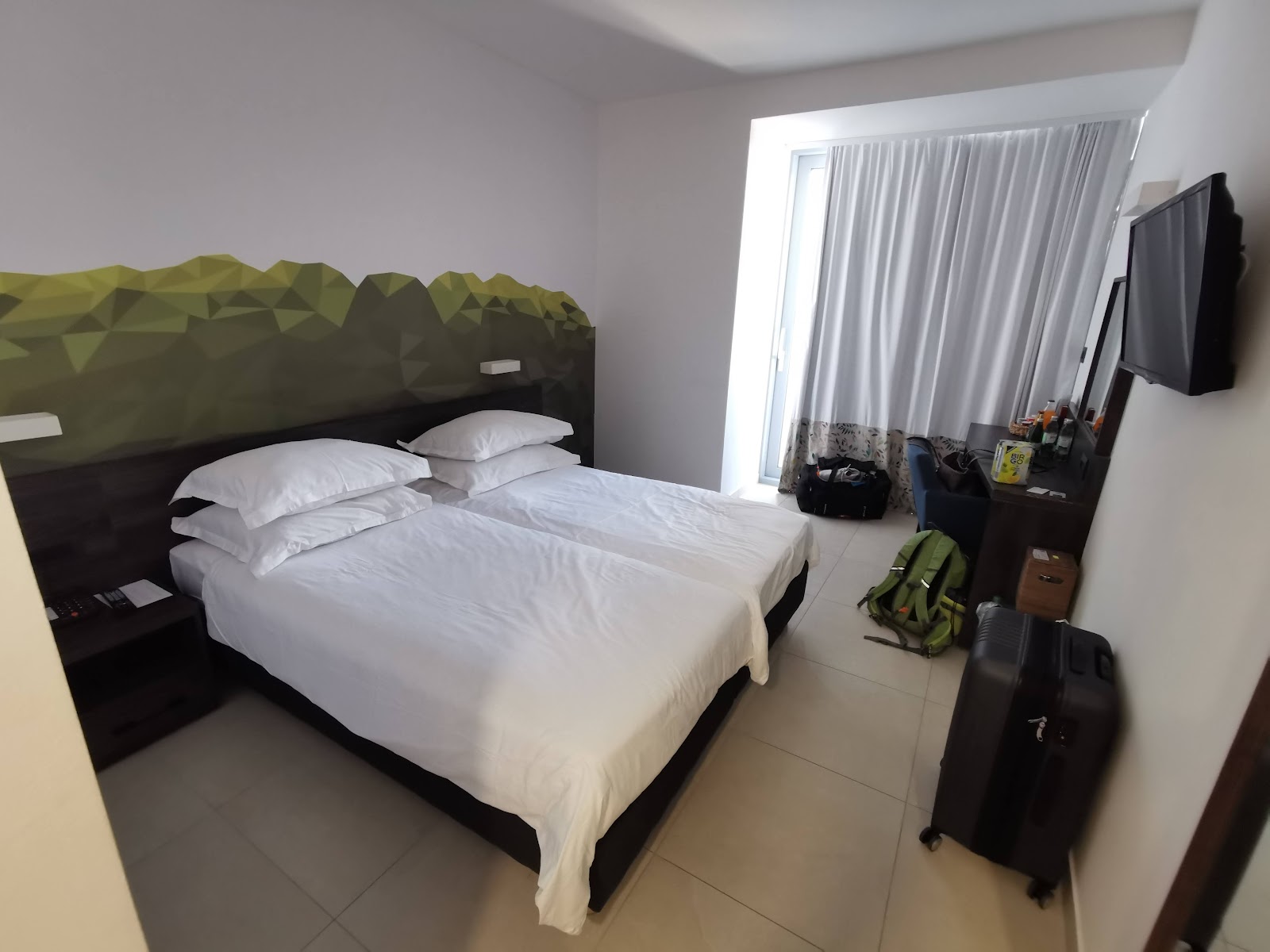 Hotel room with two beds, wooden desk with mirror, and glass doors opening to a terrace overlooking the sea and tiled roofs.