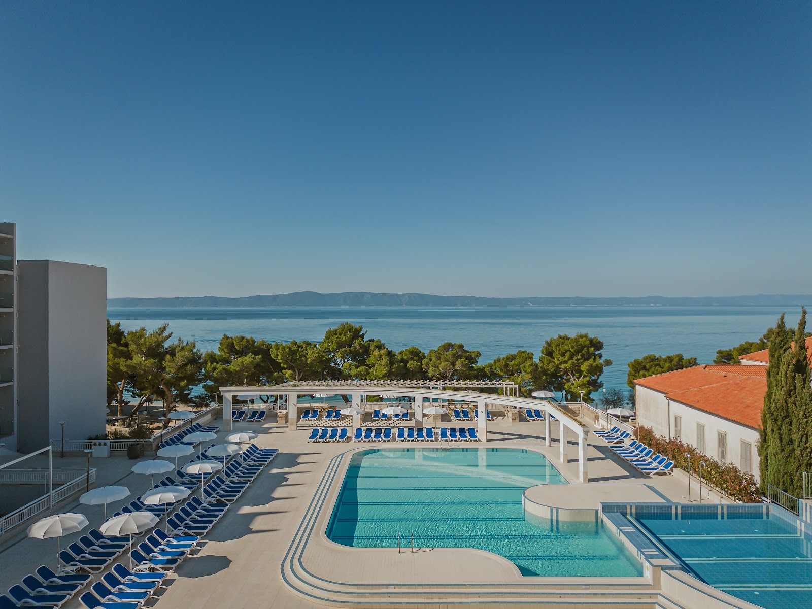 Resort pool deck with blue loungers and white umbrellas overlooking the Adriatic Sea and distant mountains under clear sky.