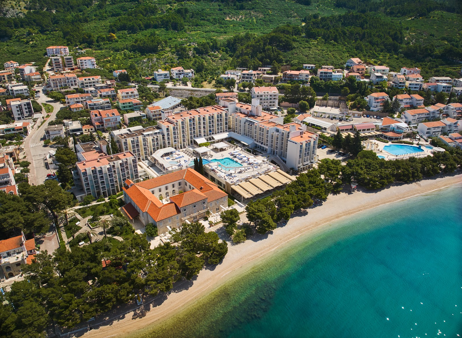 Bluesun Hotel Alga sprawls across a beachfront property surrounded by green hills, with multiple modern buildings, pools, and a sandy beach meeting turquoise water.
