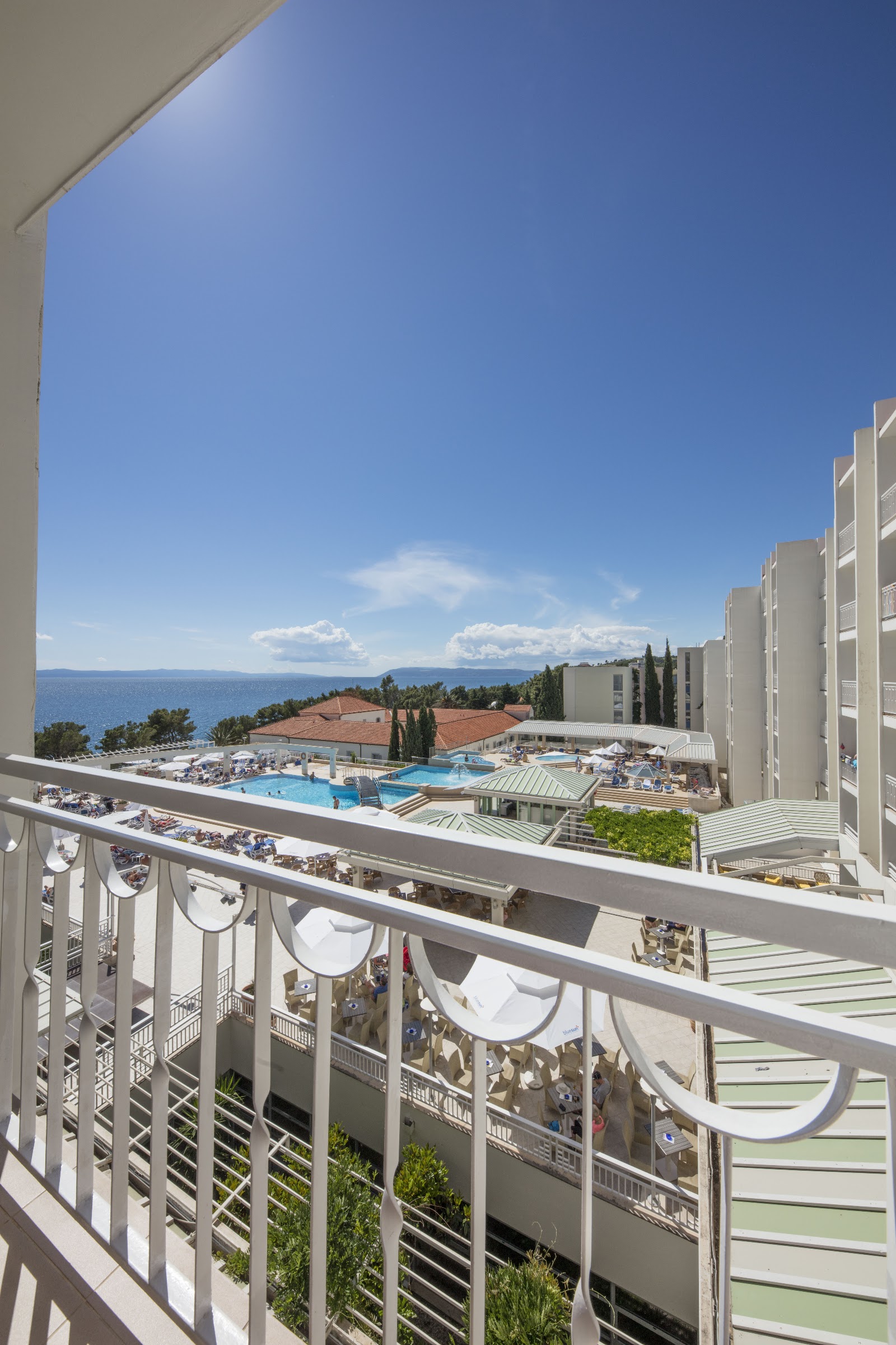 Aerial view of Bluesun Hotel Alga with multiple pools, blue loungers, and Mediterranean Sea beyond sandy beach and pine trees.
